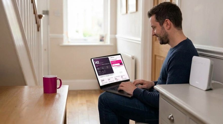 A man sits on a hall chair in a UK home with a laptop on his knees beside the broadband router on the hall table, the laptop screen showing the Pulse speed test result from BroadbandSwitch.uk, 142 Mbps, with a pink mug on the table opposite.