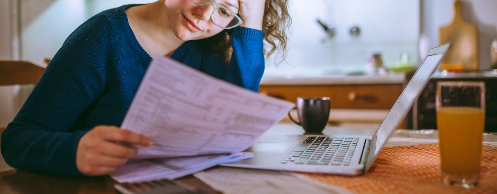 A person at a kitchen table going through household letters beside an open laptop.