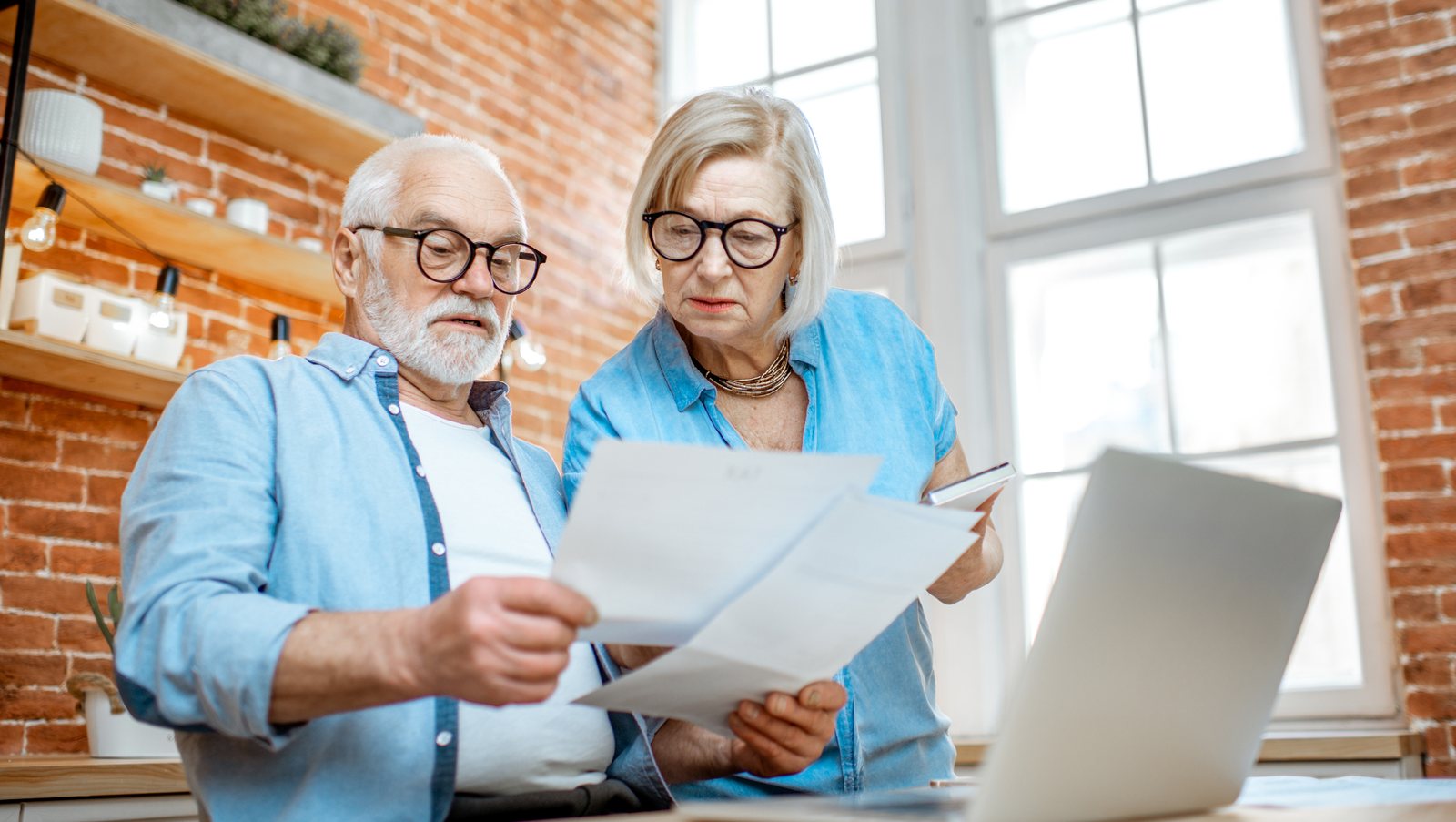 An older couple at home reading household bills together beside an open laptop.