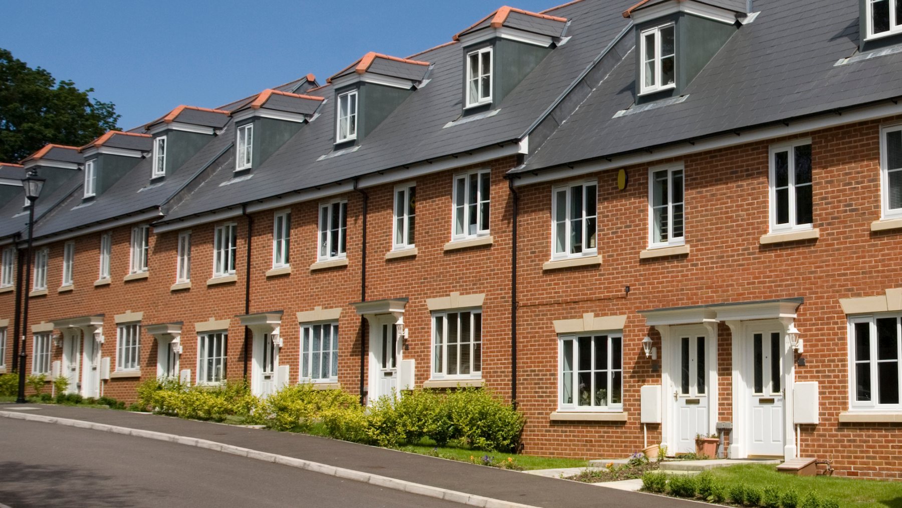 A row of modern red-brick terraced houses on a quiet UK residential street in sunshine.