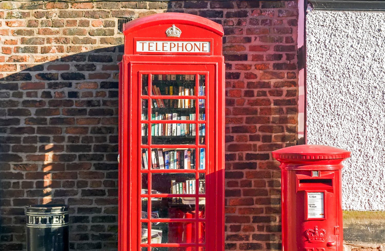 Red telephone box filled with bookshelves serving as a community book exchange, beside a red Royal Mail post box