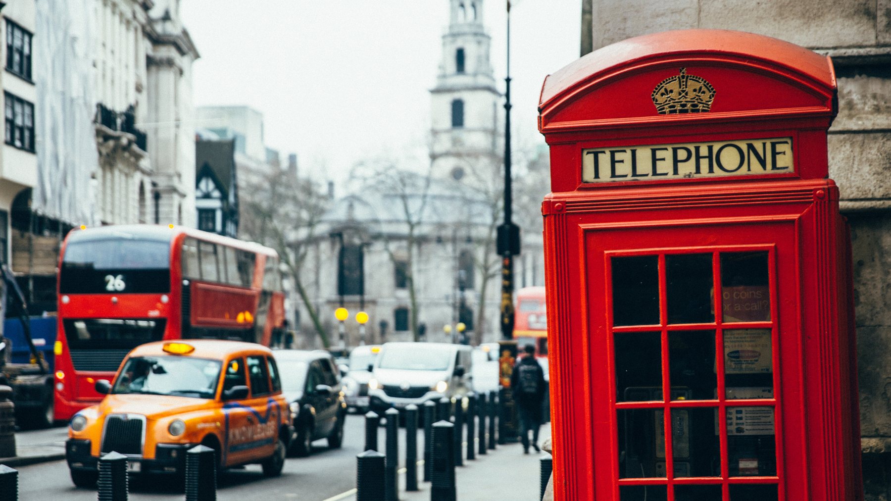 Red telephone box in the foreground of a London street with a number 26 double decker bus, a black cab and a church spire behind