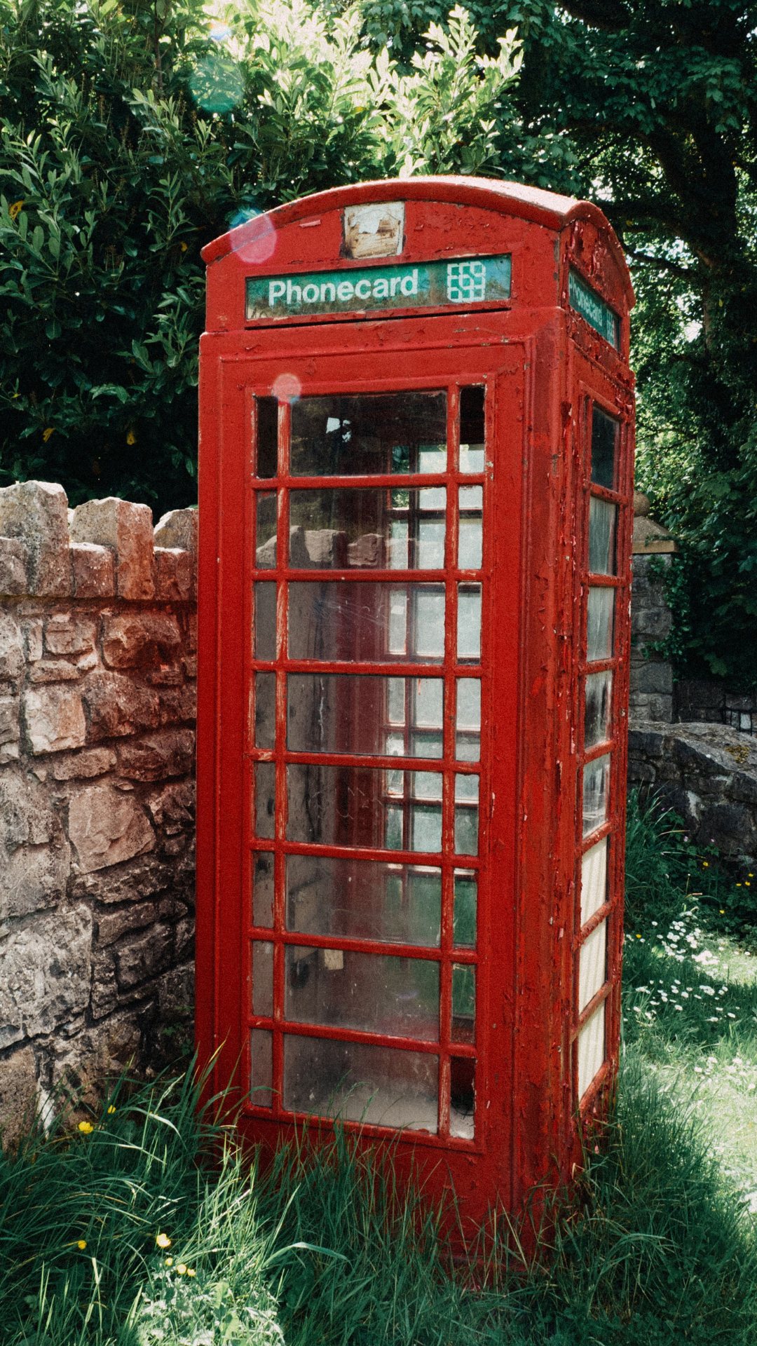 Weathered red telephone box with peeling paint and a green Phonecard sign beside a stone wall